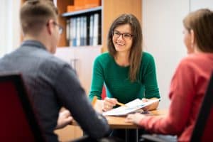 Fachhochschule Dortmund Foto einer Mitarbeiterin der Studienberatung, die mit zwei Studierenden an einem Tisch spricht. Sie hält ihnen einen Zettel hin und zeigt mit einem Stift darauf.