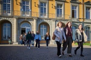 Studierende laufen von dem Hauptgebäude der Universität Bonn zum Hofgarten. Sie lachen und die tiefstehende Sonne scheint Ihnen auf die Gesichter.